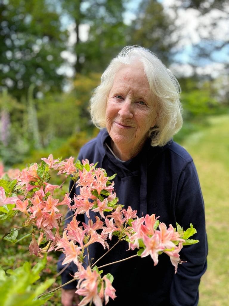 Elderly woman outdoors with pink azalea flowers, sunny day, natural setting.