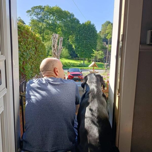 A man and a black dog sitting on a doorstep, looking out at a sunny garden scene.