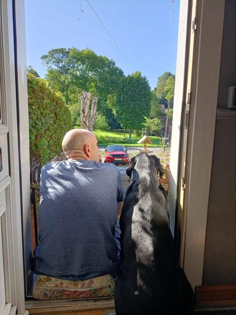 A man and a black dog sitting on a doorstep, looking out at a sunny garden scene.