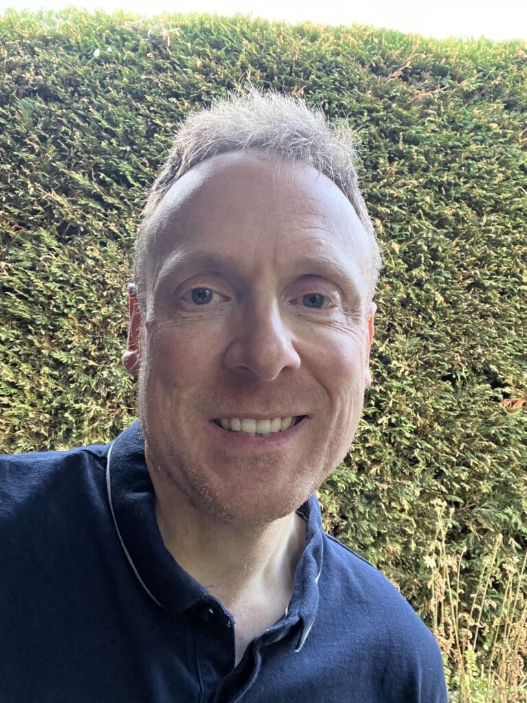 Smiling man outdoors with a hedge background, wearing a navy shirt.
