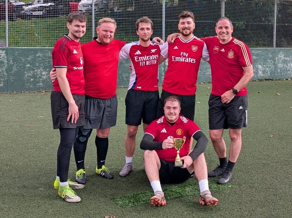Group of five men in football kits celebrating on a pitch with a trophy.